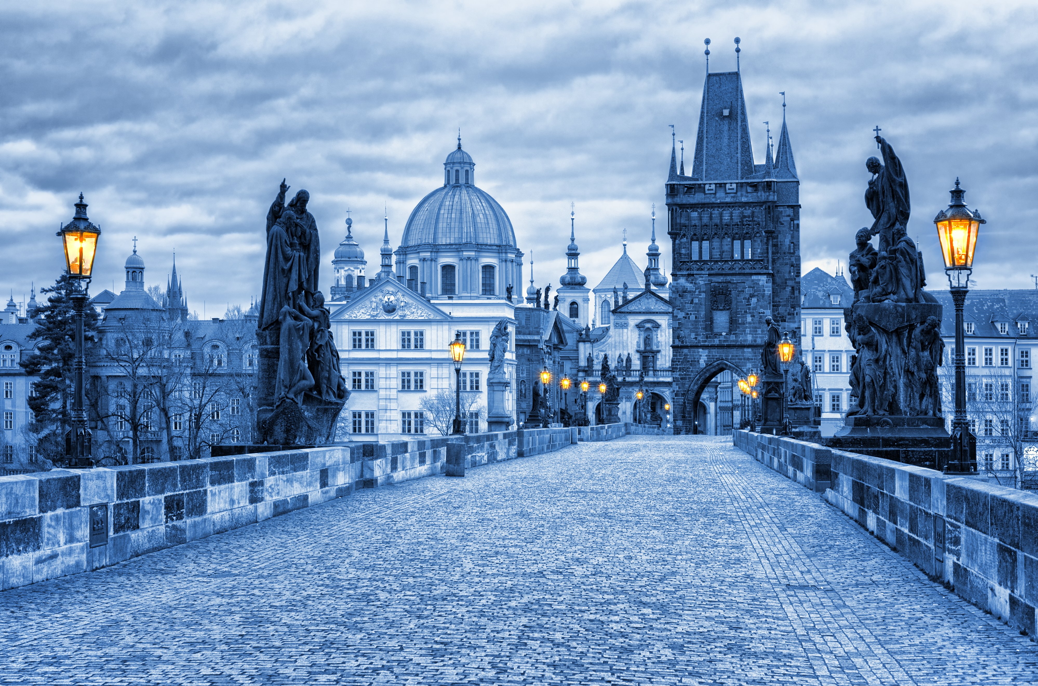 Charles Bridge in Prague at dusk, its statues and glowing lamps leading toward the Old Town tower