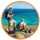 Two hikers with backpacks resting on rocks beside the bright blue sea